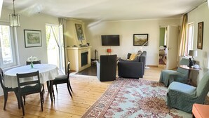 A living room with a fireplace, a television, and a dining area with chairs and a table. - Top location on the lake - Charming yellow bungalow with terrace & lake view (Neuruppin)