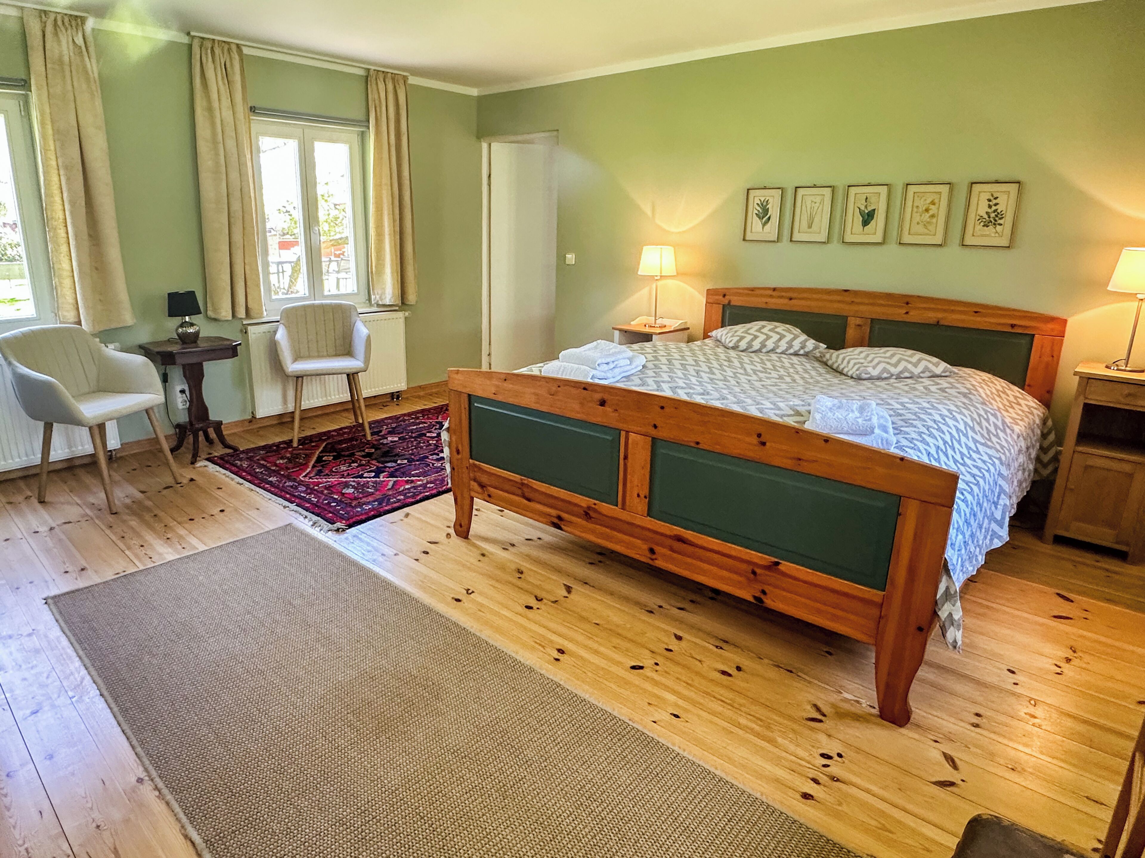 A bedroom with a wooden bed, two chairs, a small table, a radiator, and framed botanical prints on the wall.