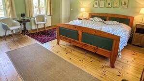 A bedroom with a wooden bed, two chairs, a small table, a radiator, and framed botanical prints on the wall. - Top location on the lake - Charming yellow bungalow with terrace & lake view (Neuruppin)