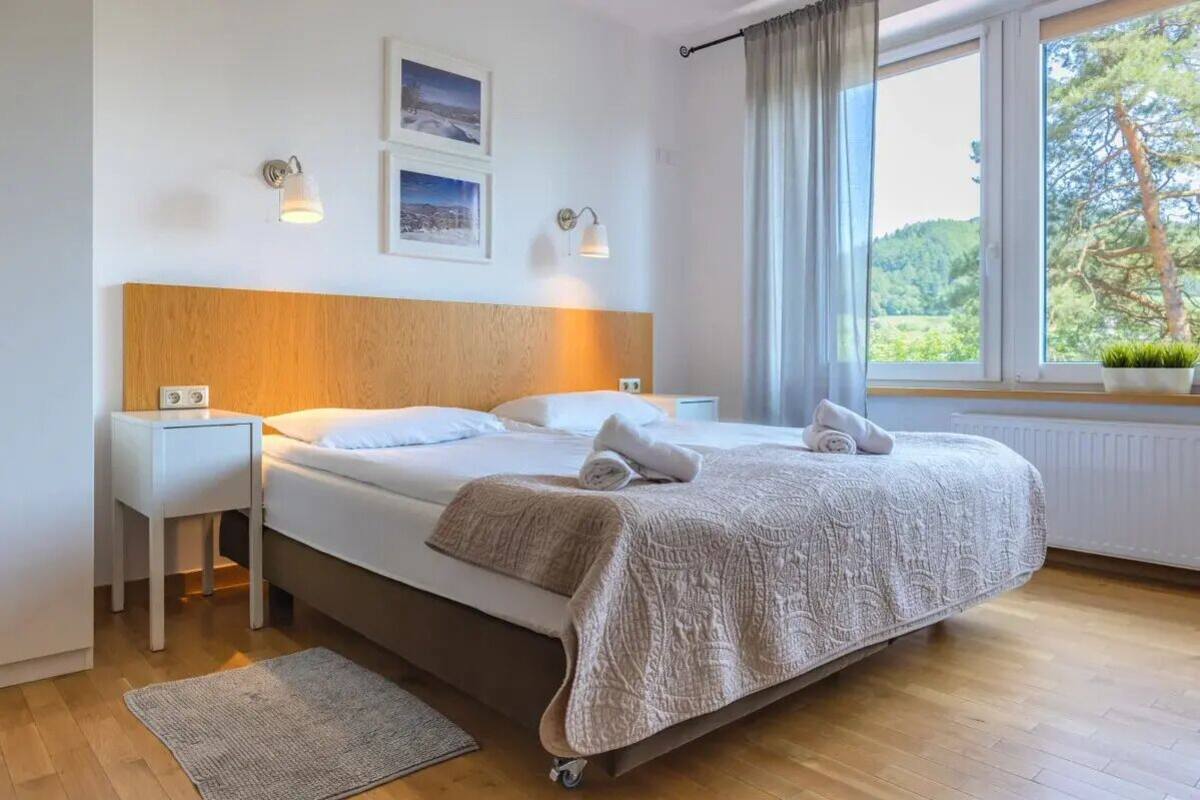 Une chambre à coucher avec une tête de lit en bois, un lit recouvert d’une couverture à motifs, deux tables de chevet, une lampe de chevet, une fenêtre donnant sur un paysage verdoyant et deux appliques murales.