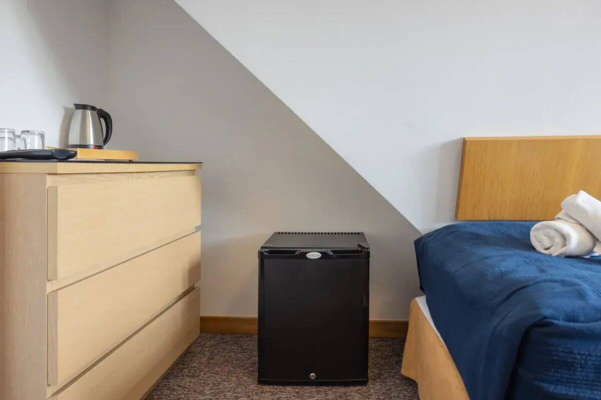 Une chambre à coucher avec un lit, une commode en bois, une poubelle noire et une bouilloire posée sur la commode.