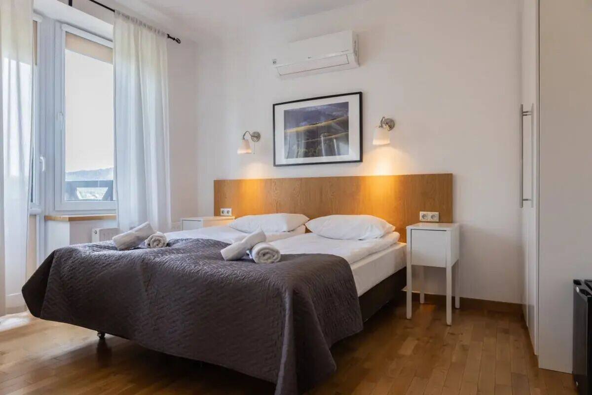 Une chambre d’hôtel avec un lit, des tables de chevet, une fenêtre avec des rideaux, un appareil de climatisation et un tableau encadré au mur.