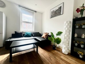A modern living room with a sofa, a coffee table, a potted plant, a white brick wall, and a framed picture. - 2BR Apartment in the heart of Streatham High road (London)