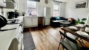 Een kleine keuken met een fornuis, spoelbak en kastjes, een eethoek met een tafel en stoelen, en een bank.