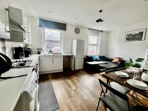 A compact kitchen with a stove, sink, and cabinets, a dining area with a table and chairs, and a sofa. - 2BR Apartment in the heart of Streatham High road (London)