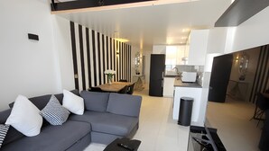 Ein modernes Wohnzimmer mit einem Ecksofa, einem Couchtisch und einem Fernseher. Im Hintergrund ist eine Küchenzeile mit einem Kühlschrank und einer Spüle zu sehen.