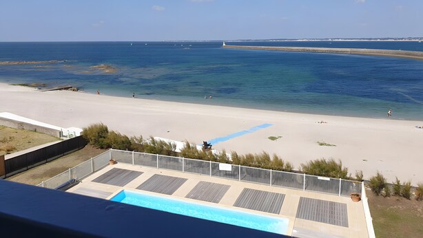 Blick auf einen Strand mit Poolbereich, ein Leuchtturm in der Ferne und einem klaren Himmel.