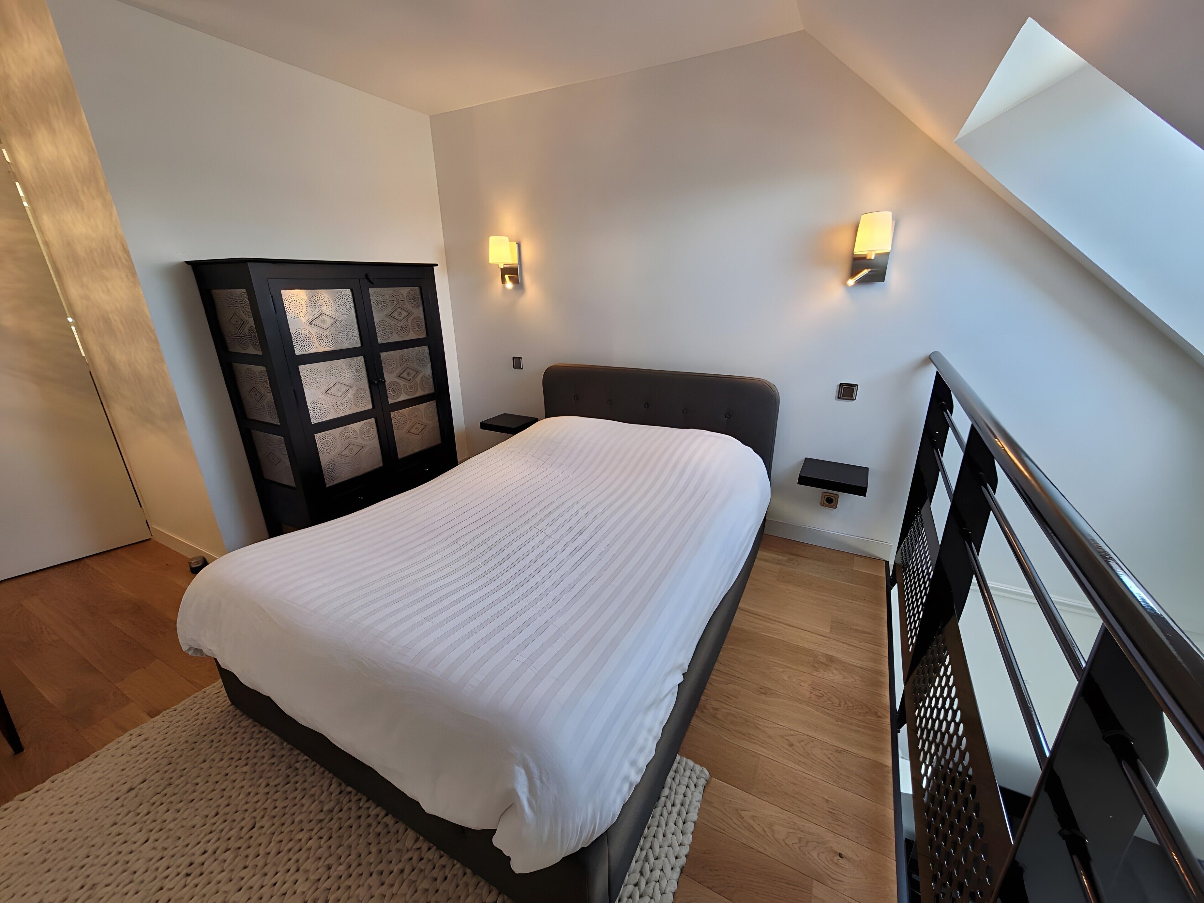 Une chambre à coucher comprenant un lit, une table de chevet, une lampe fixée au mur, une armoire et un escalier.