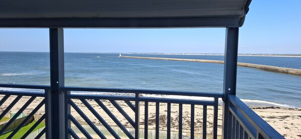 Vue sur la plage depuis un balcon couvert doté d’une balustrade, avec une plage de sable et une digue en arrière-plan.