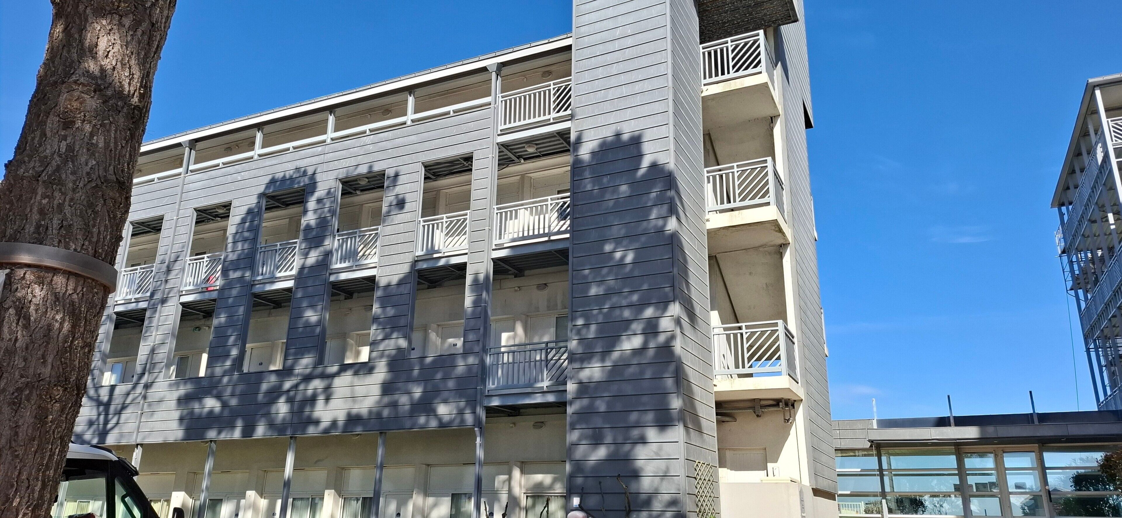 Un immeuble à plusieurs étages doté de balcons, un arbre au premier plan et un ciel bleu dégagé.