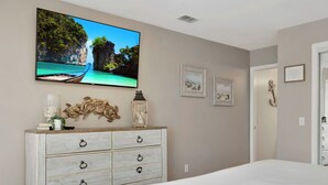 A bedroom with a bed, a dresser, a television mounted on the wall, and framed pictures on the wall. - 🏖️Steps to Beach | Great Outdoor Spaces | Perfect for Families | Beach Dreams☀️ (St. Joe Beach)