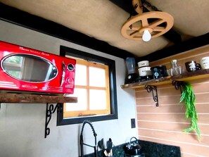 A kitchen with a red microwave, a window, a black kettle, and a wooden shelf with kitchen items.