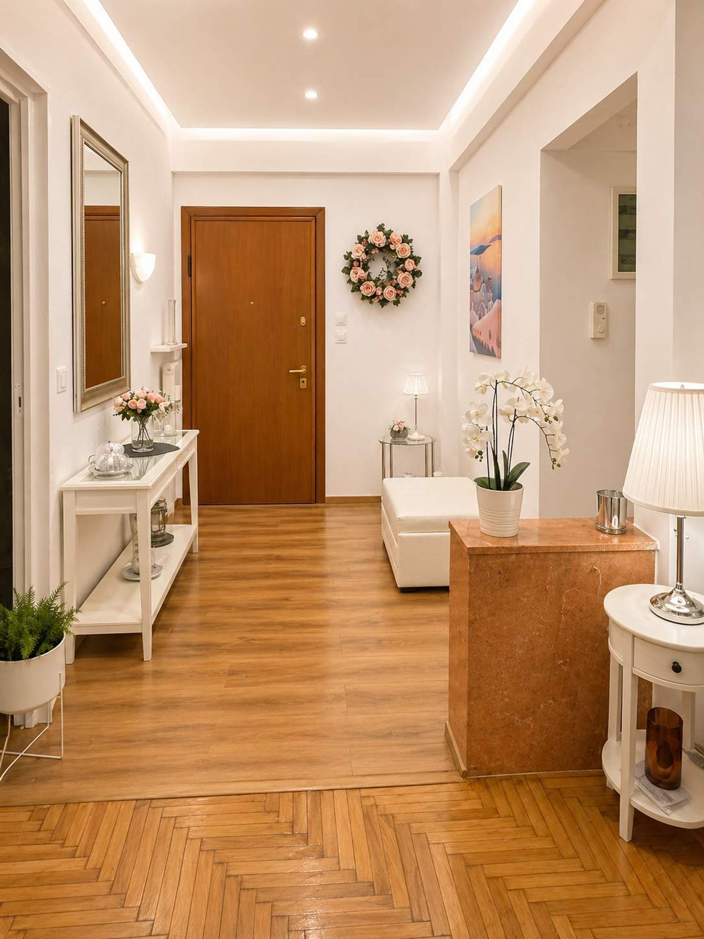 A hallway with a wooden floor, a white console table with a vase of flowers, a white bench, a wooden door, and a wall-mounted painting.
