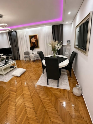 Ein modernes Wohnzimmer mit Fischgrätparkett, einem runden Esstisch mit Stühlen, einem Fernseher und einem Gemälde an der Wand.