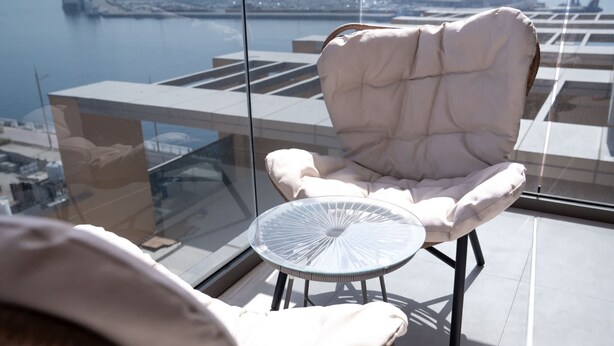A high-rise balcony with a glass railing, a cushioned chair, a small round table, and a view of a body of water. - Free Housekeeping Visits During Your Stay - Stayshort - Stunning 1 Bed in Maritime City That Sleeps 2 (Dubai)