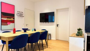 Een moderne eetruimte met een houten tafel, blauwe stoelen, een groot abstract schilderij, een aan de muur bevestigde televisie en een deur met een sleutelgat.