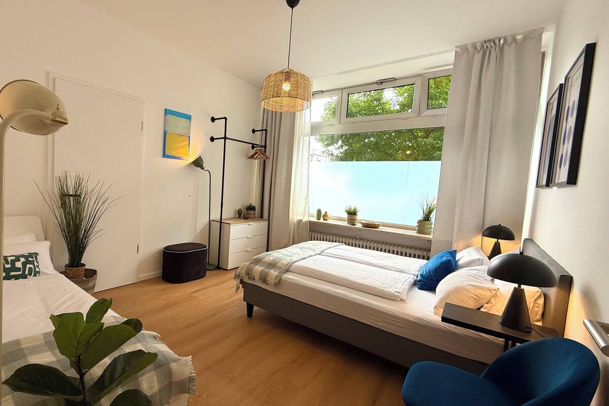 Quarto com cama, mesa de cabeceira, luminária, planta em vaso e janela com cortinas.