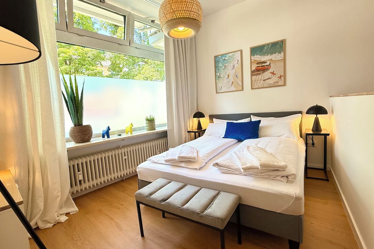 Um quarto bem arrumado com uma cama, luminárias de cabeceira, um banquinho, uma janela com cortinas e um radiador.