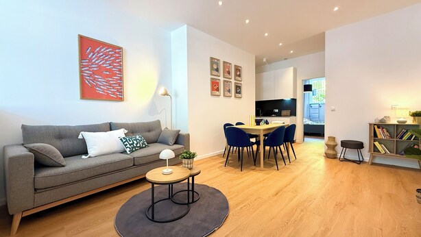 Een moderne woonkamer met een grijze bank, een ronde salontafel en een eethoek met blauwe stoelen.