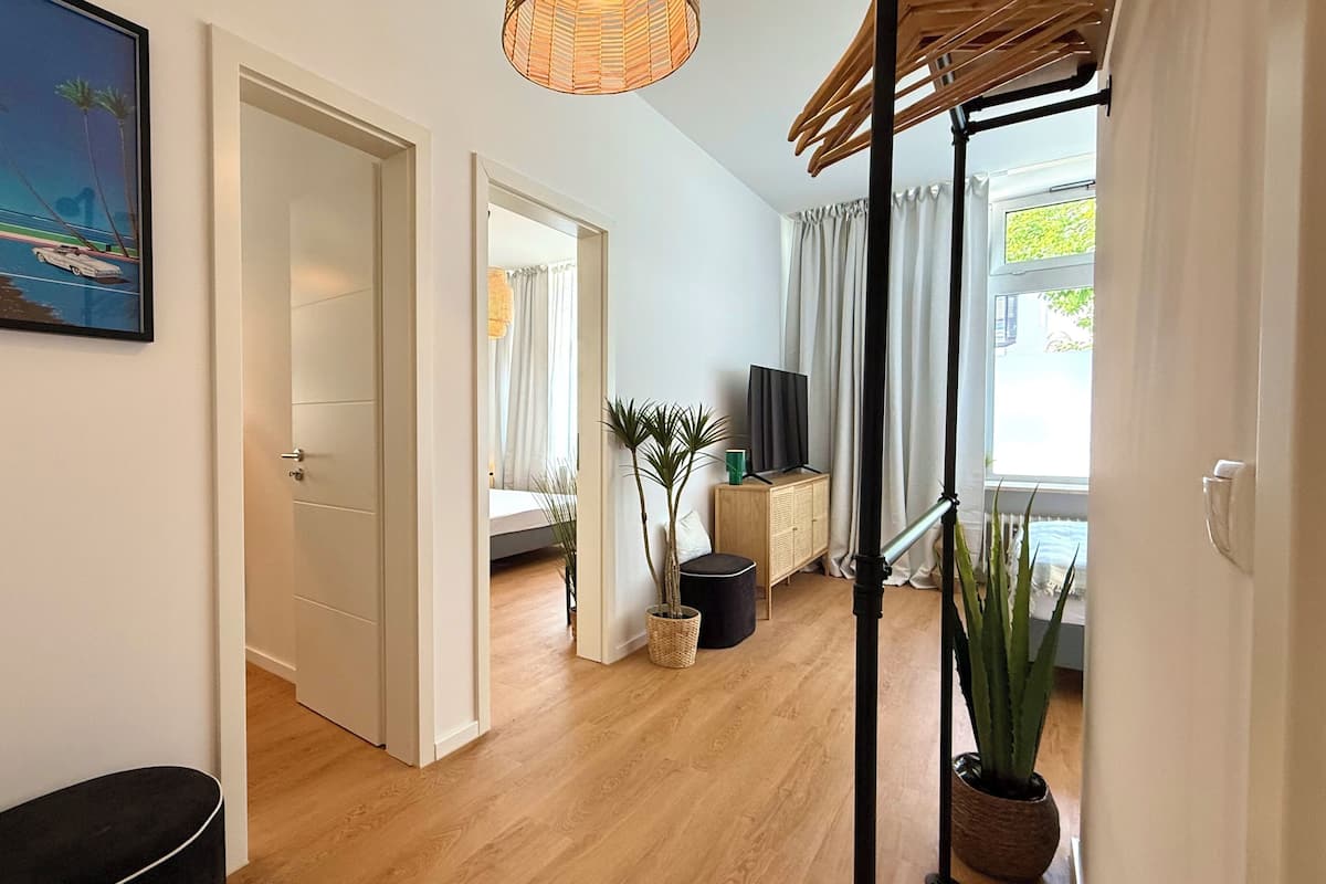 Um corredor com uma porta que dá para um quarto, uma televisão sobre um móvel e um vaso de plantas.
