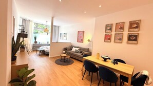 Een moderne woonkamer met een bank, een eettafel met stoelen, een klein rond tafeltje met een lamp en een groot raam met gordijnen.