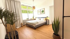 Slaapkamer met een bed, een raam, een radiator, een nachtkastje, een lamp en twee potplanten.