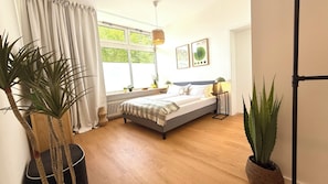 A bedroom with a bed, a window, a radiator, a bedside table, a lamp, and two potted plants. - BeckHomes Stylish Central Apartments (Munich)