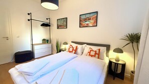 Una cama bien hecha con ropa blanca y almohadas naranjas, una mesita de noche con una lámpara, una lámpara de pie y dos obras de arte enmarcadas en la pared.