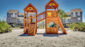 Una estructura de juegos de madera en una playa arenosa, con edificios residenciales altos de fondo.