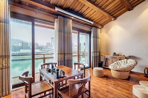 Quarto com mesa de madeira, cadeiras, sofá e vista para um rio e edifícios.