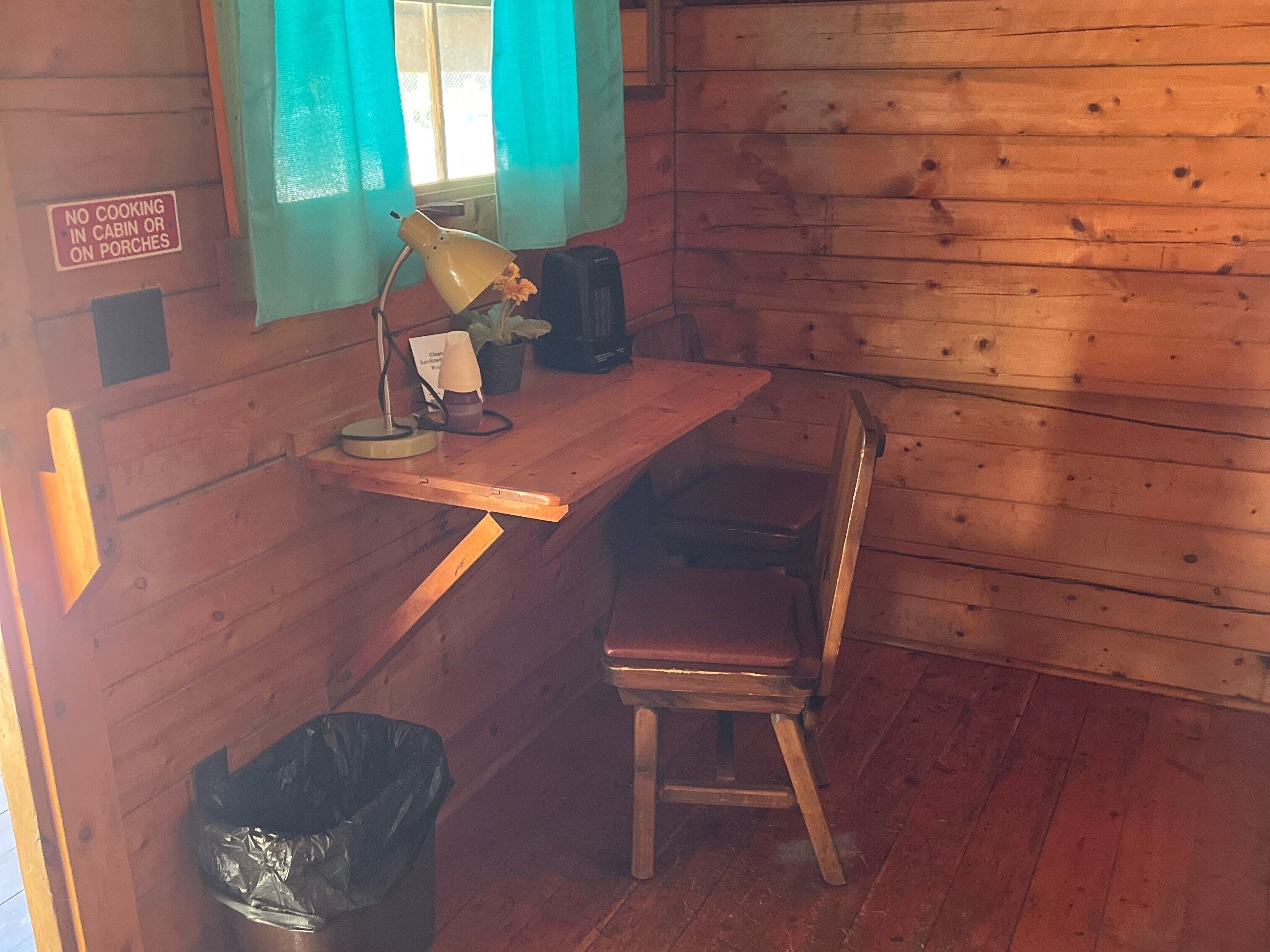 A cabin interior with a wooden desk, a chair, a lamp, a trash bin, and a window with curtains.