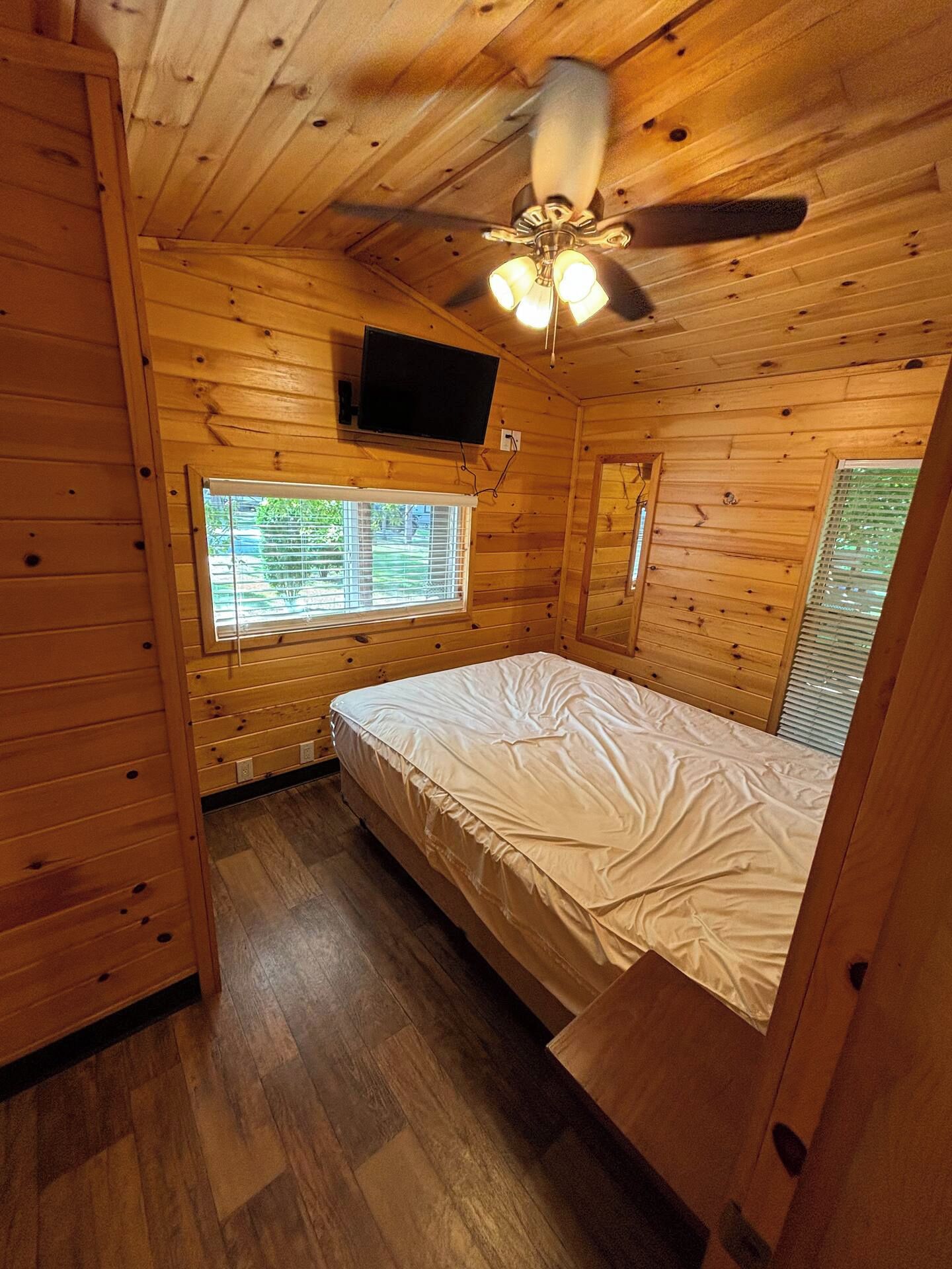 A wooden cabin bedroom with a bed, a ceiling fan, a TV, and windows.