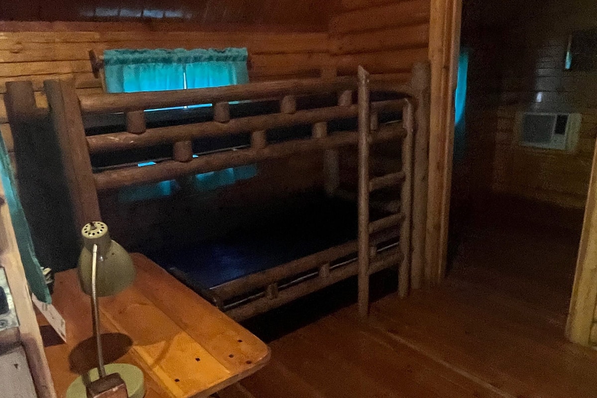 A wooden bunk bed with a ladder, a small table with a lamp, and a window with curtains.