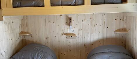 A room with two beds, wooden walls, a ceiling light, and a shelf with pillows.