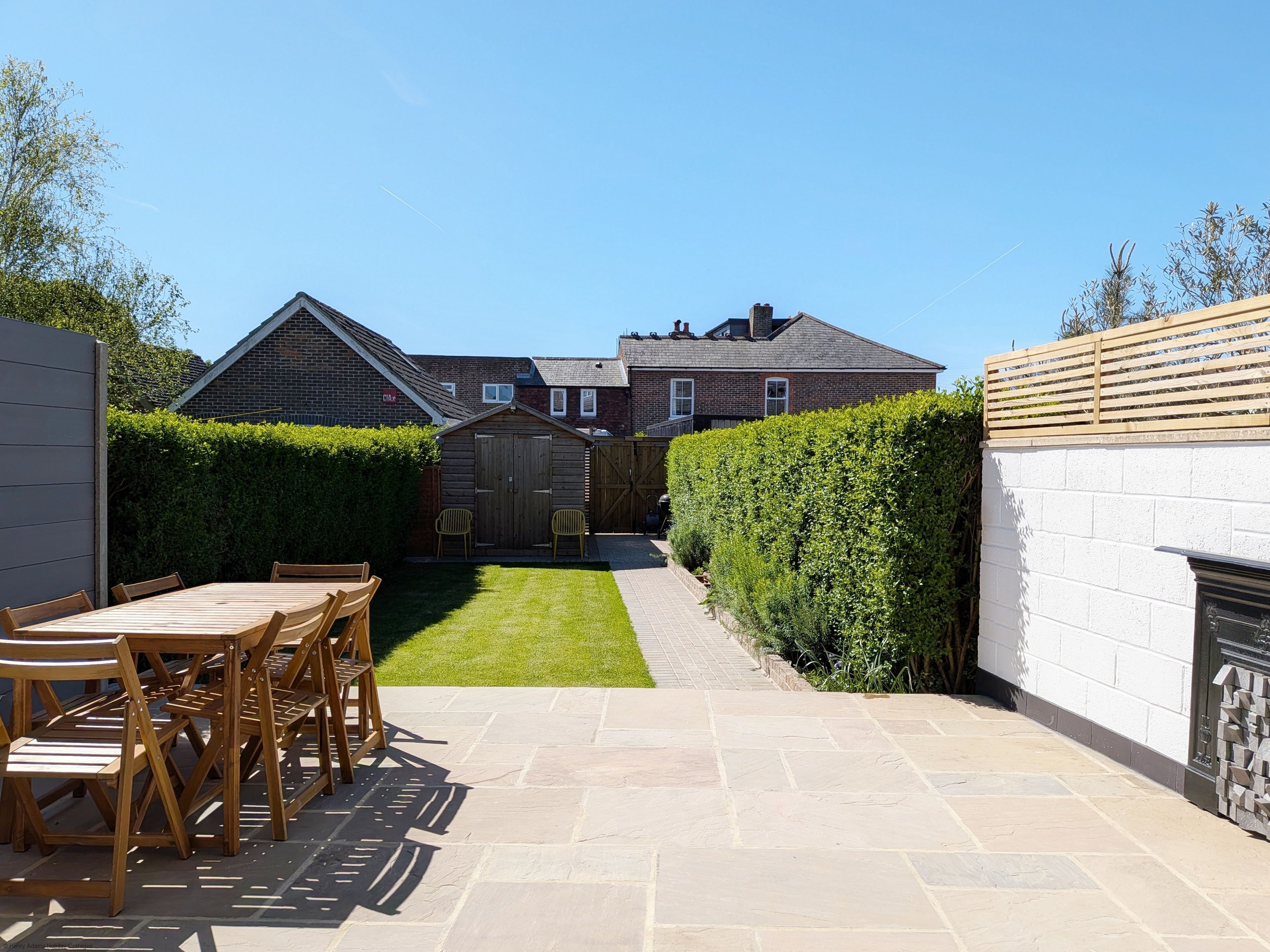 A patio area with a wooden table and chairs, a small shed, and a hedge.