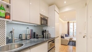 A modern kitchen with a stainless steel sink, a microwave, and a coffee machine. There is a seating area with a sofa and a table, and a view of the city through a window.