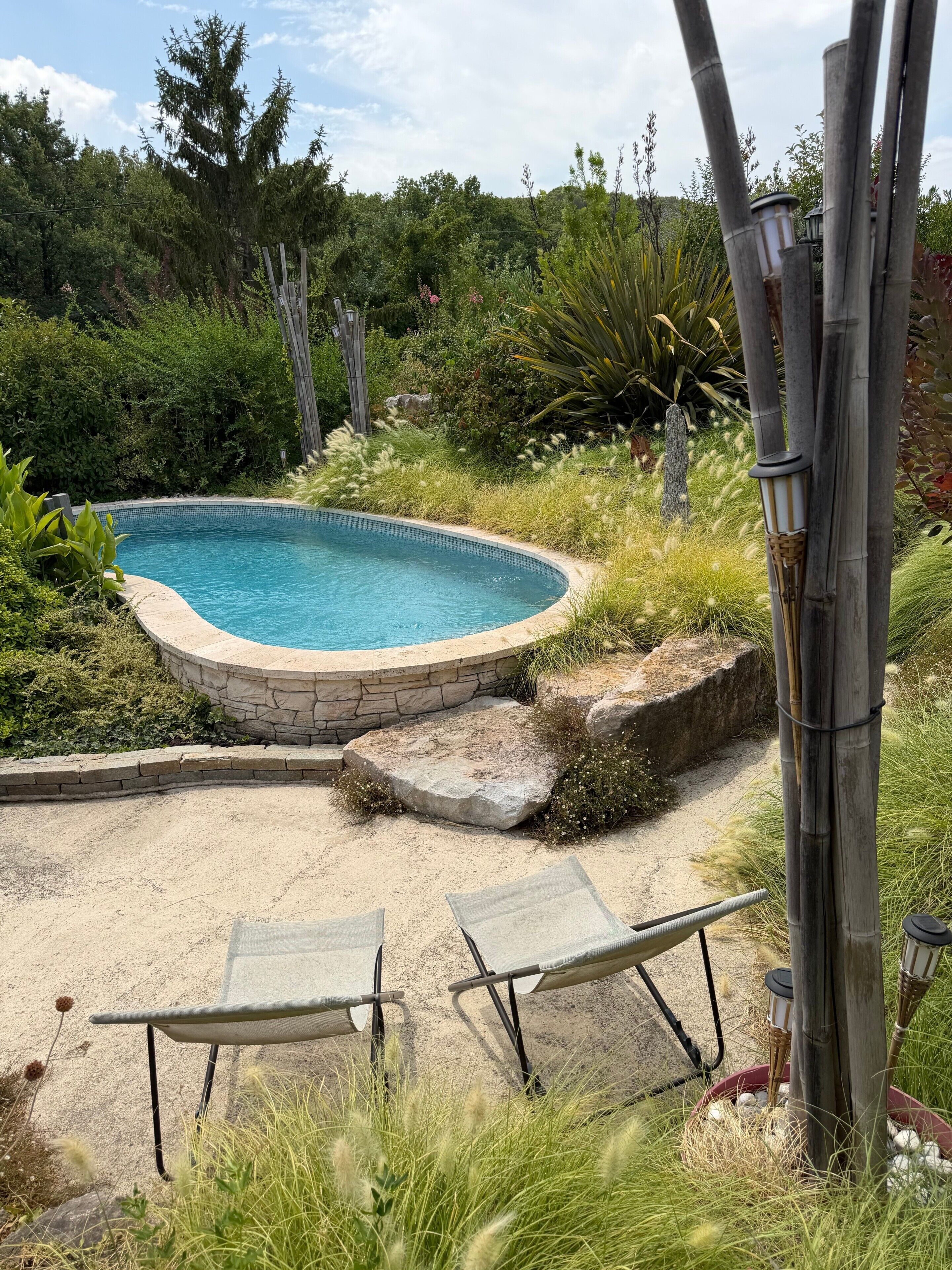 Ein Schwimmbecken mit Steinrand, umgeben von einem Garten mit verschiedenen Pflanzen und einer Bambuskonstruktion.