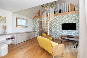 A spacious living area with a wooden staircase, a yellow sofa, a dining table, and a TV mounted on the wall.