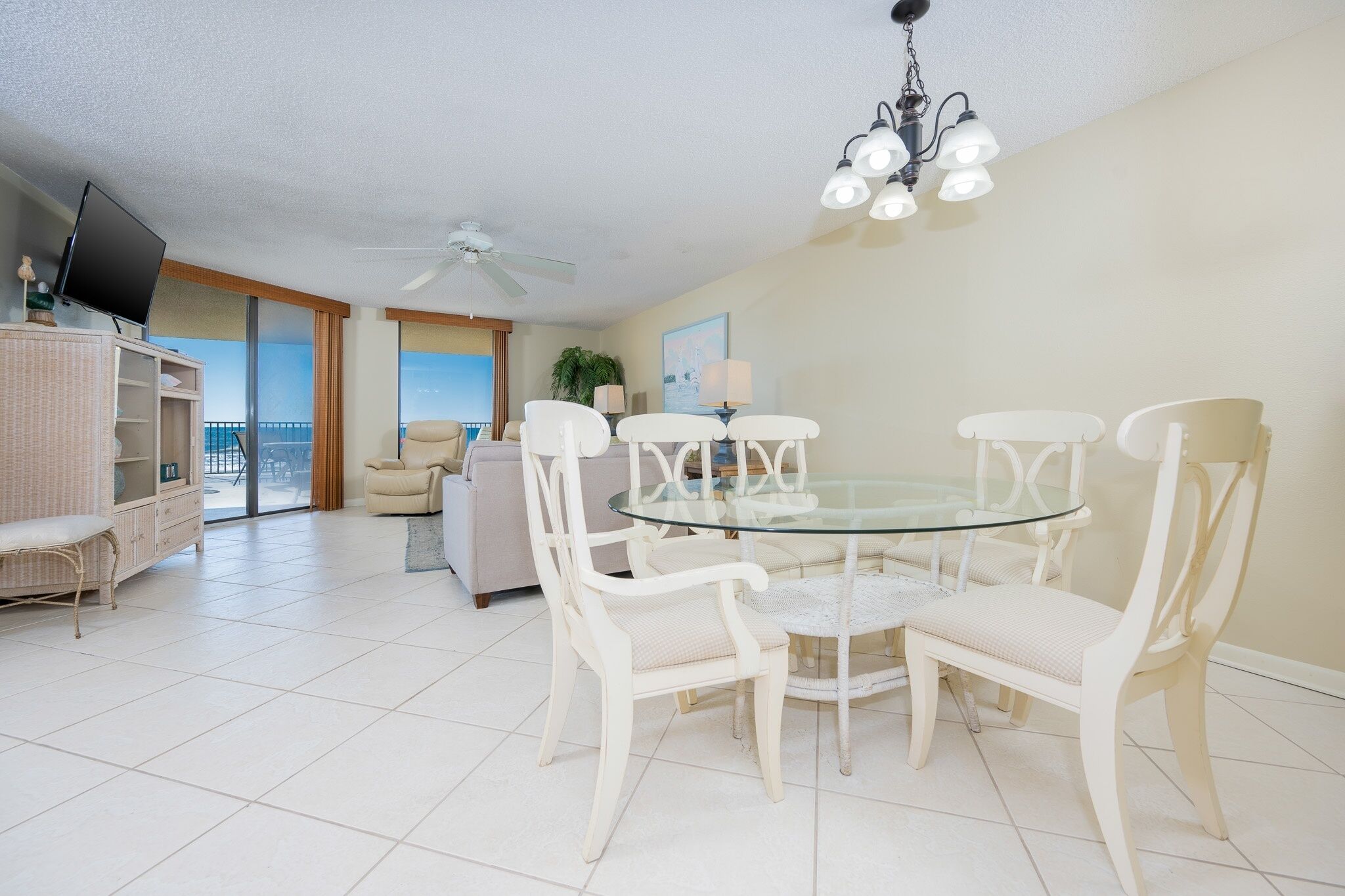 A dining area with a glass table and white chairs, a ceiling fan, and a view through large windows.