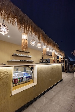 Un espace bar avec un comptoir, un présentoir pour les boissons, des luminaires suspendus et un toit de chaume.