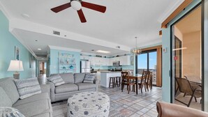 A spacious living area with a sofa, ottoman, and dining table, a kitchen in the background, and a balcony with outdoor seating. - Phoenix V 1117 3 Bedroom Condo (Orange Beach)