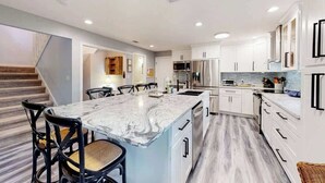 A modern kitchen with a marble island, white cabinetry, and a staircase leading to another room. - 3BR Beach Getaway w Balcony (Cape Canaveral)