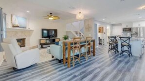 A modern living room with a fireplace, a dining area with a wooden table and chairs, a kitchen with white cabinets, and a staircase leading to another level. - 3BR Beach Getaway w Balcony (Cape Canaveral)