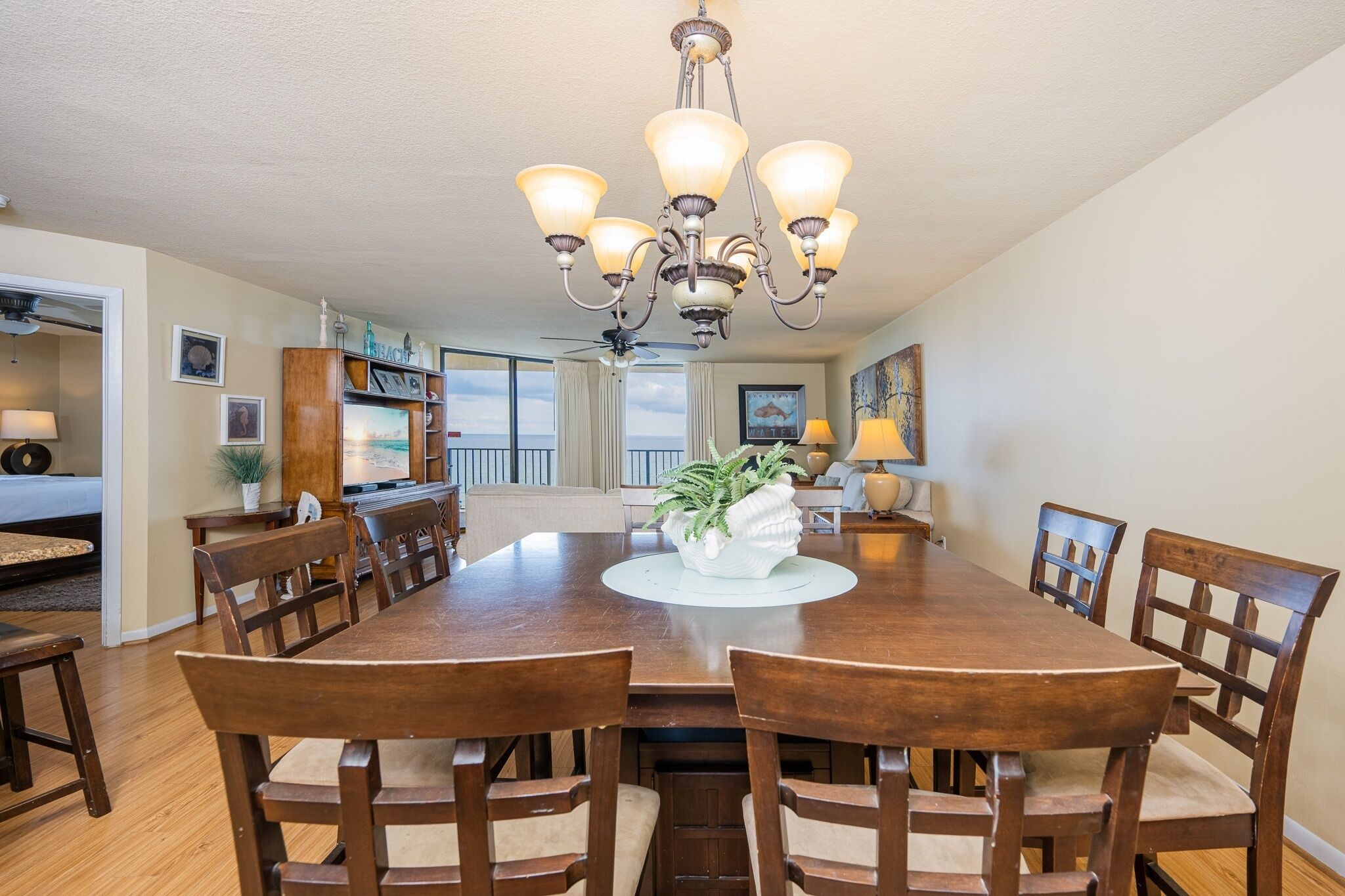 A dining area with a large wooden table, six chairs, a chandelier, and a view of the ocean.