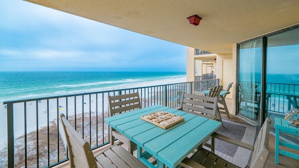 A balcony with a view of the beach and ocean, wooden furniture, and a table with a tray of snacks. - Phoenix V 708 3 Bedroom Condo (Orange Beach)