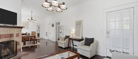 A living room with a fireplace, a television, a chandelier, and a dining area with a table and chairs.