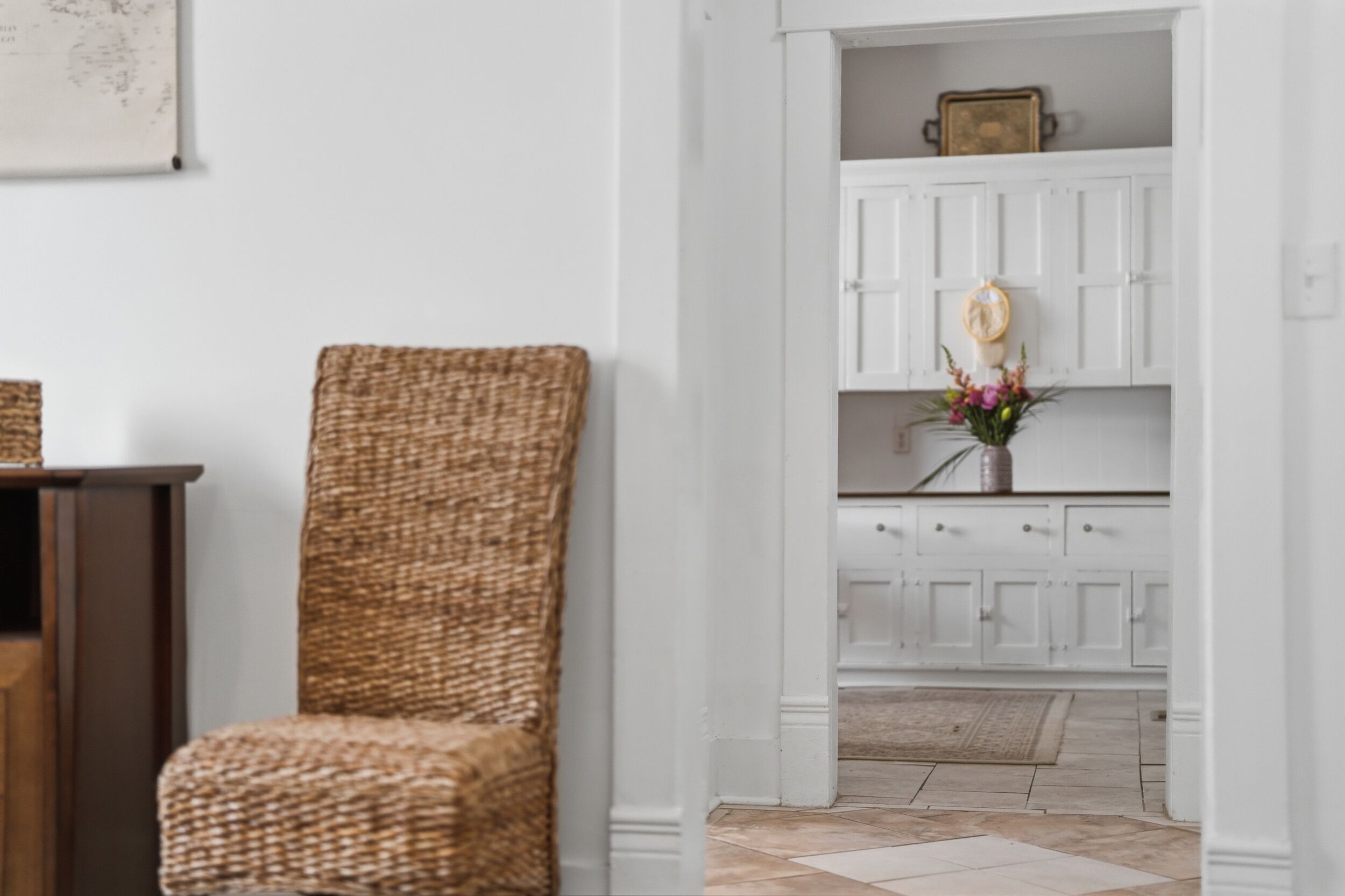Una silla de mimbre, un armario blanco con cajones y un jarrón con flores.