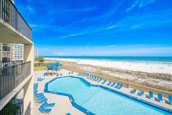 A pool with blue chairs, a beach view, and a building with balconies. - Phoenix V 209 2 Bedroom Condo (Orange Beach)