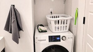 A compact laundry room with a washing machine, a drying rack, a hanging towel, and a broom. - Cozy Studio Downtown Detroit Steps from Wayne State University (Detroit)