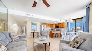 A modern living room with a round coffee table, grey sofas, and a kitchen area with wooden cabinets and a dining set. - Phoenix V 817 3 Bedroom Condo (Orange Beach)
